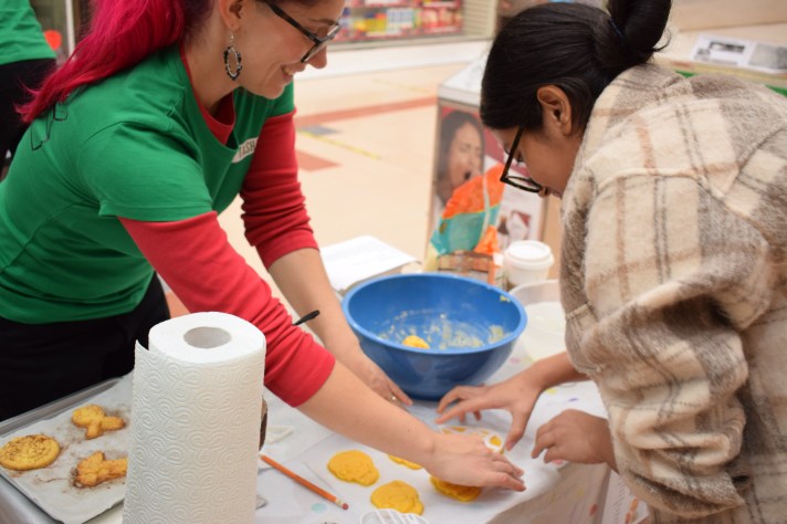 A woman wearing the branded Coffin Tales Roadshow t-shirt helps a young woman to cut out skull-shaped biscuits with a cookie cutter.