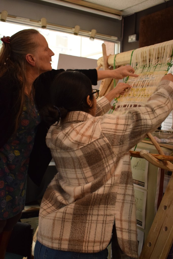 A young woman and an older woman weave cloth tape on a standing loom.