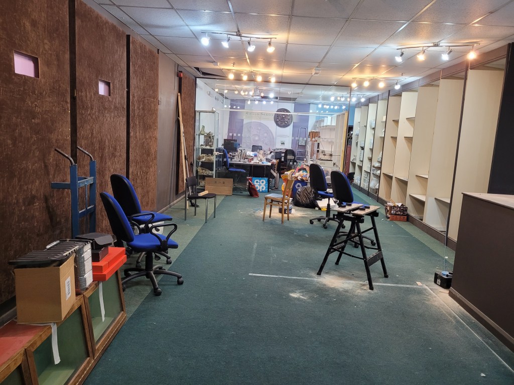 The same space mostly empty, but for some office chairs and workbench. Towards the back there is a glass wall that opens into the new lab space which contains a desk and shelves.