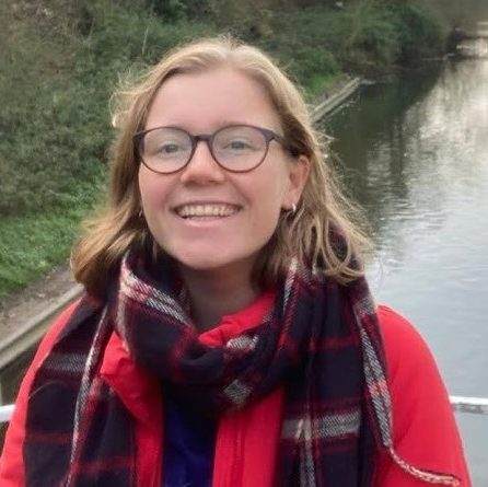Headshot of Emma in front of a river, a white person with light brown hair and glasses.