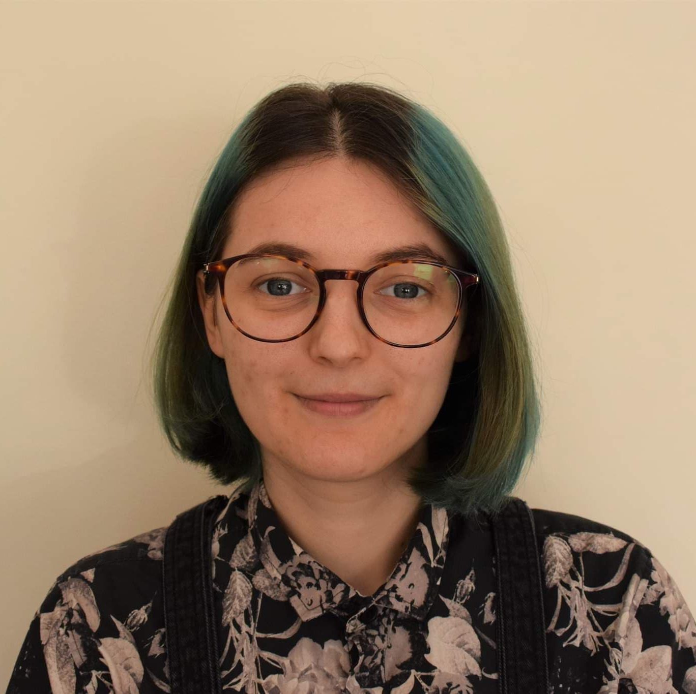 Headshot of Morgan, a white person in their mid-20s. They have green shoulder length hair and tortoiseshell glasses.