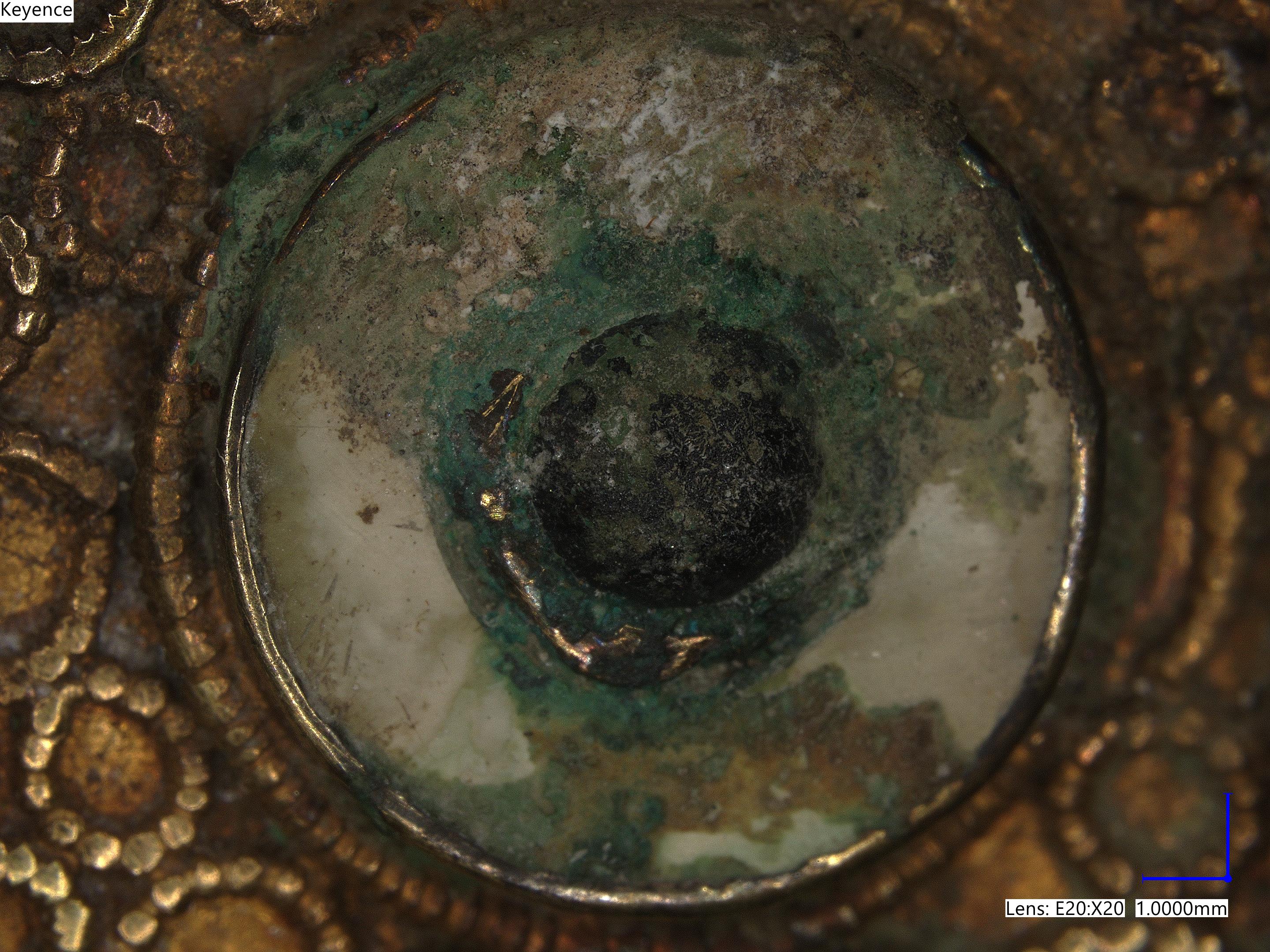 Microscope close-up of a brooch, showing tiny details of flaking metals and circular patterns of gold bumps.