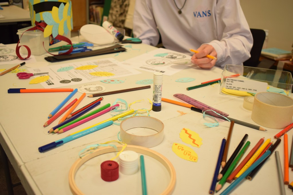 Messy table with craft supplies - pens, pencils, tapes, glue, card - and worksheets about Roman coins