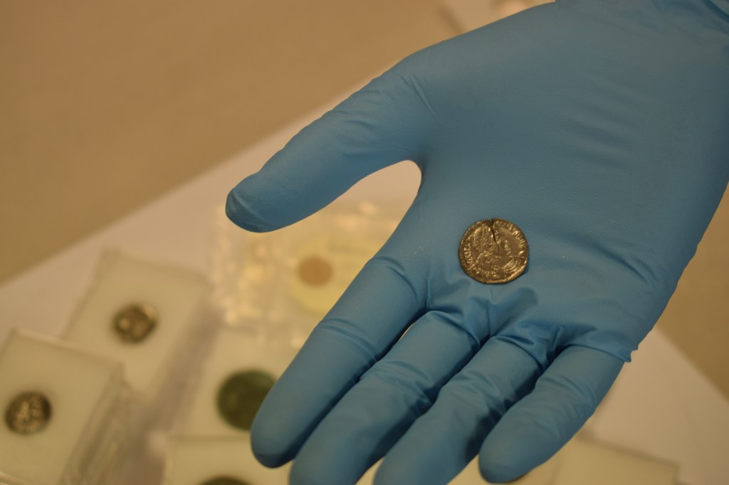 Shiny silver coin, about the size of a 10p coin, with emperor's head and Latin text on. The person holding it is wearing blue conservation gloves